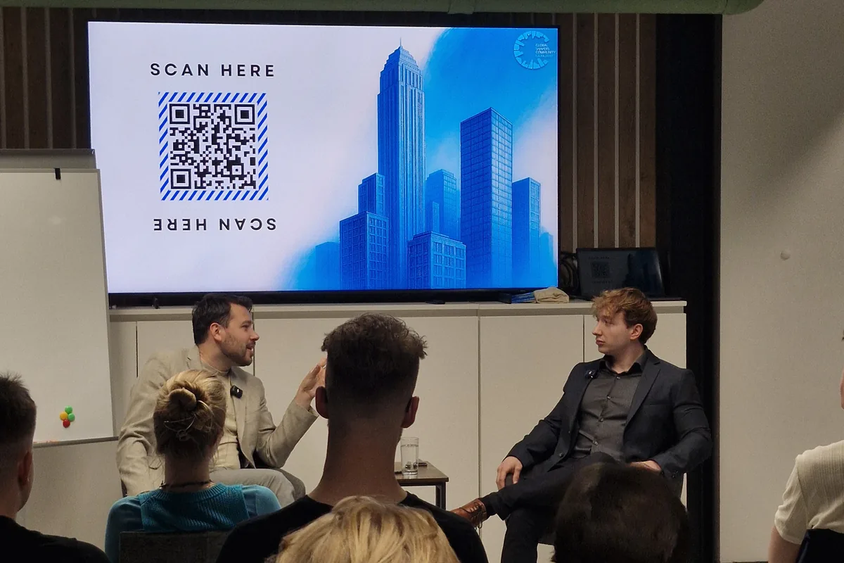 Two speakers seated in front of a screen showing a QR code and a blue skyline illustration at Impact Hub Ljubljana, the audience watching from the rows in front.
