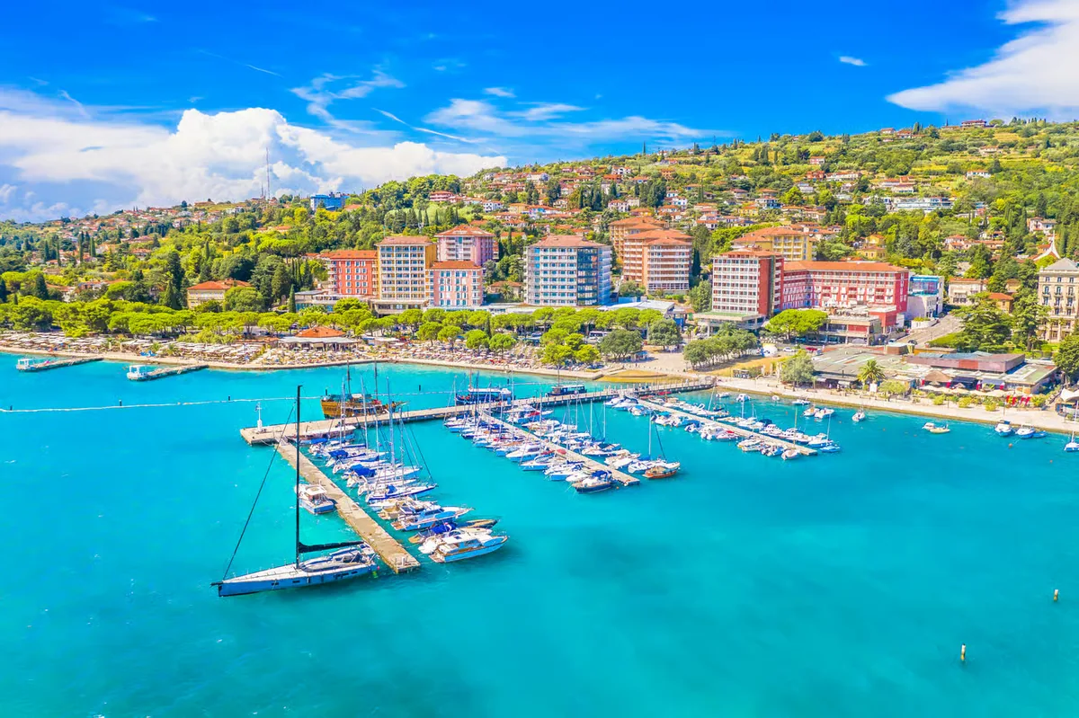 Aerial view of the Portorož marina with sailboats at the pontoons, turquoise sea, beachfront hotels and the green hillside behind.