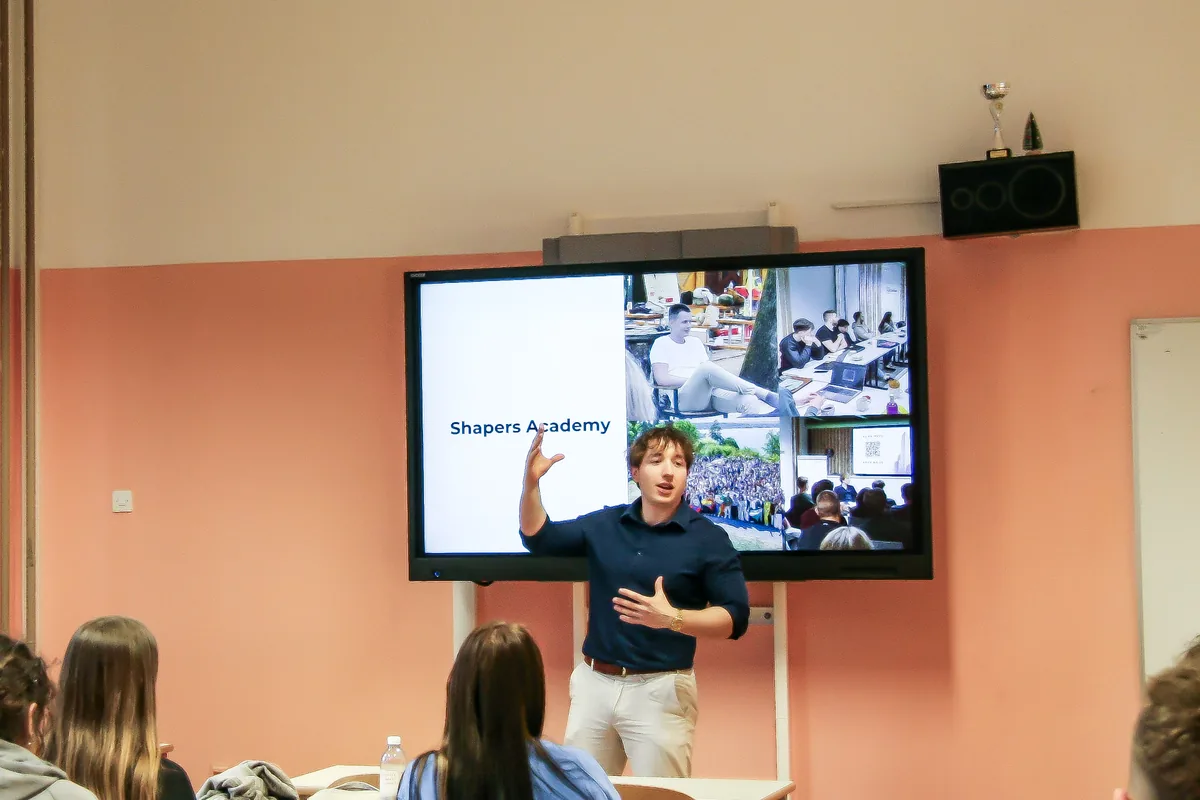 The presenter gesturing towards a slide titled “Shapers Academy” with a grid of programme photos, the students following from their desks.