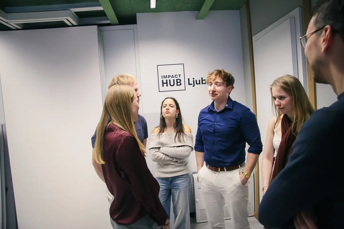 Five visitors standing in a circle and talking after the talk, in front of the Impact Hub Ljubljana wall sign.
