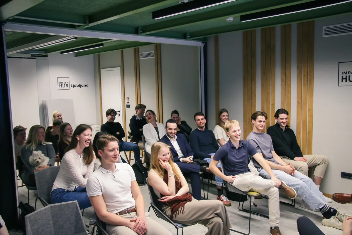 About twenty young people sitting in rows at Impact Hub Ljubljana, laughing at something said on stage, the Impact Hub Ljubljana sign on the wall behind them.