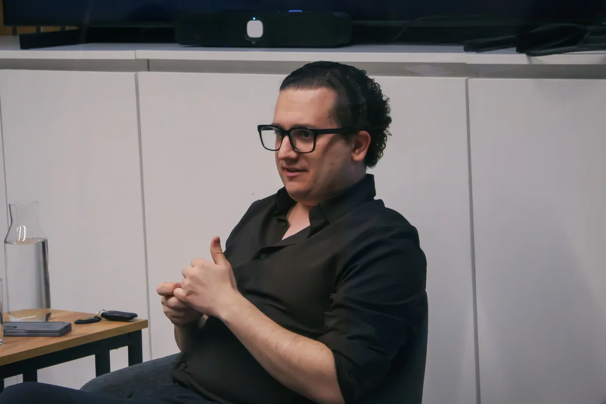 Guest Patrik Trobec, in a black shirt and heavy black glasses, answering a question from his chair on the stage at Impact Hub Ljubljana on 17 March 2026.