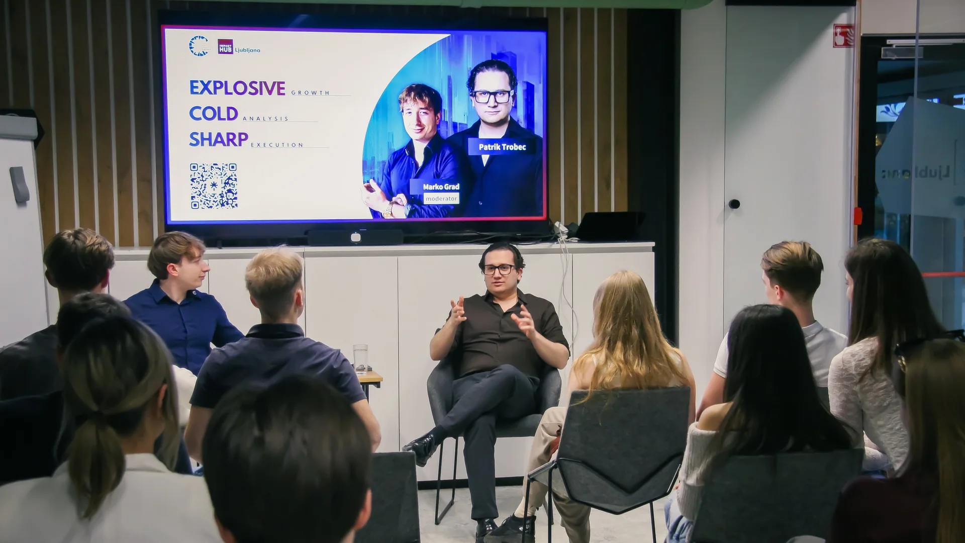 Guest Patrik Trobec speaking from a chair on the small stage at Impact Hub Ljubljana, the title slide “Explosive growth, cold analysis, sharp execution” on the screen behind him and the audience watching from the front rows.