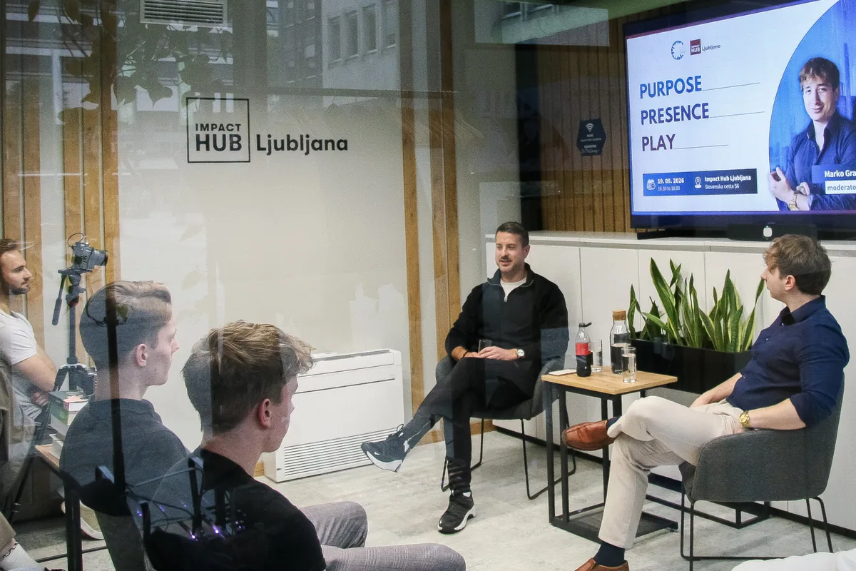 Klemen Selakovič and moderator Marko Grad seated on the stage, seen through the glass wall with the Impact Hub Ljubljana lettering, a camera on a tripod filming from the left.