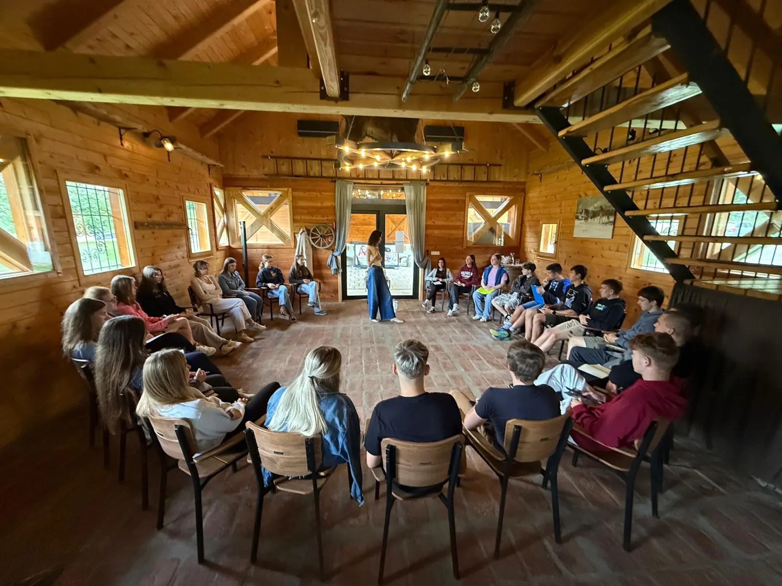 Summer school participants sitting in a circle of chairs in a wooden hall while a speaker stands in the middle