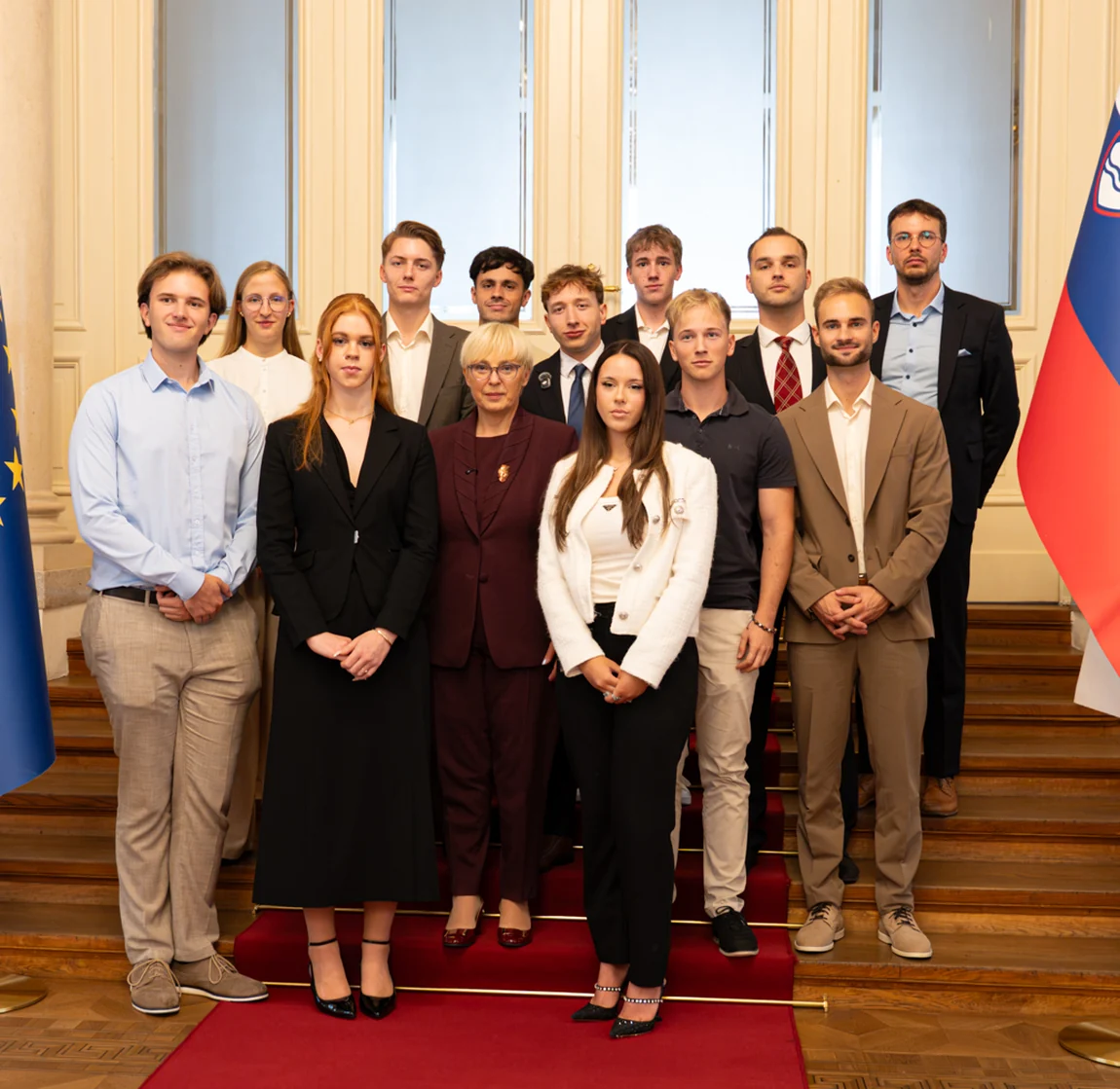 Global Shapers members in a group photograph in a formal hall, Slovenian and European flags behind them and a red carpet underfoot.